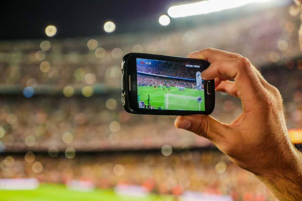 Hand holding a smartphone filming a floodlit football match from the stands