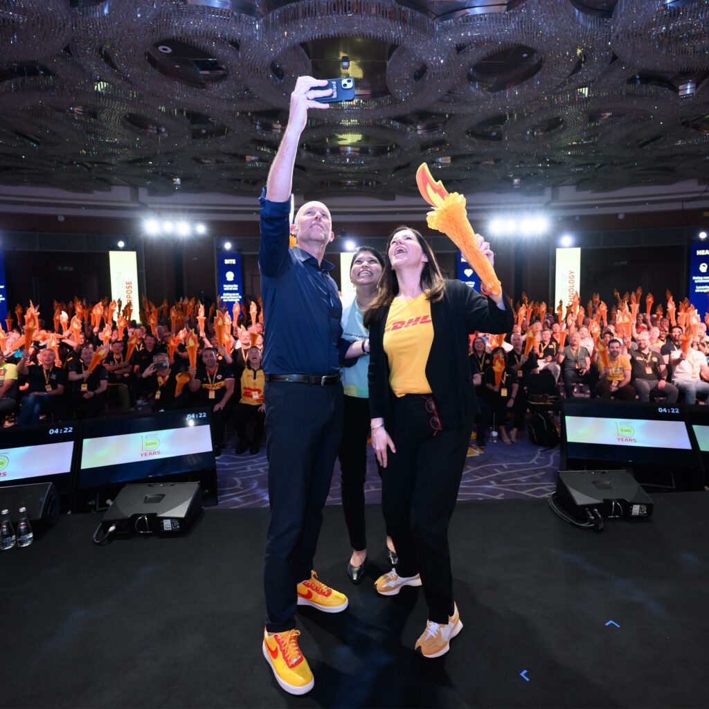 Three DHL colleagues take a selfie on stage as the audience behind them holds up hundreds of paper torches.