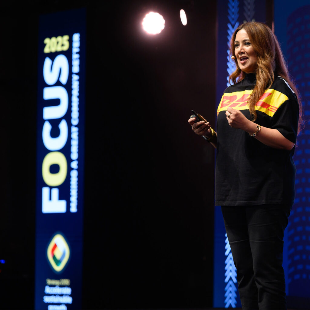 A speaker in a DHL shirt presenting on the FOCUS 2025 stage beside a tall banner carrying the event identity.