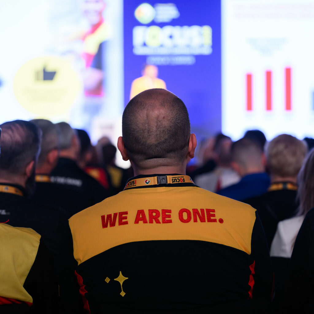 A delegate seen from behind wearing a DHL shirt embroidered with "We are one", watching a presentation.