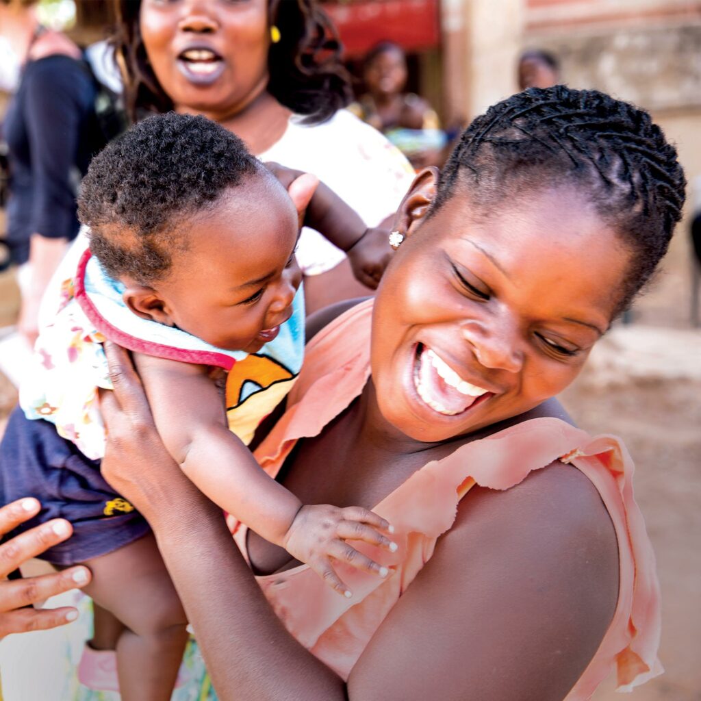 A woman laughing as she lifts a small child onto her shoulder outside a health clinic, with other mothers and babies behind them.