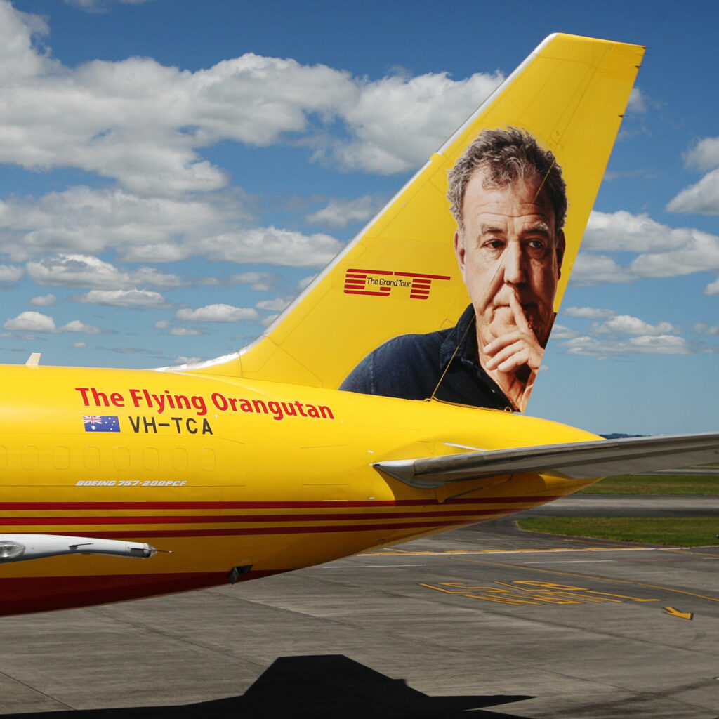 A yellow DHL Boeing 757 on the apron, its tail fin carrying a large portrait of Grand Tour presenter Jeremy Clarkson, with "The Flying Orangutan" painted along the fuselage.