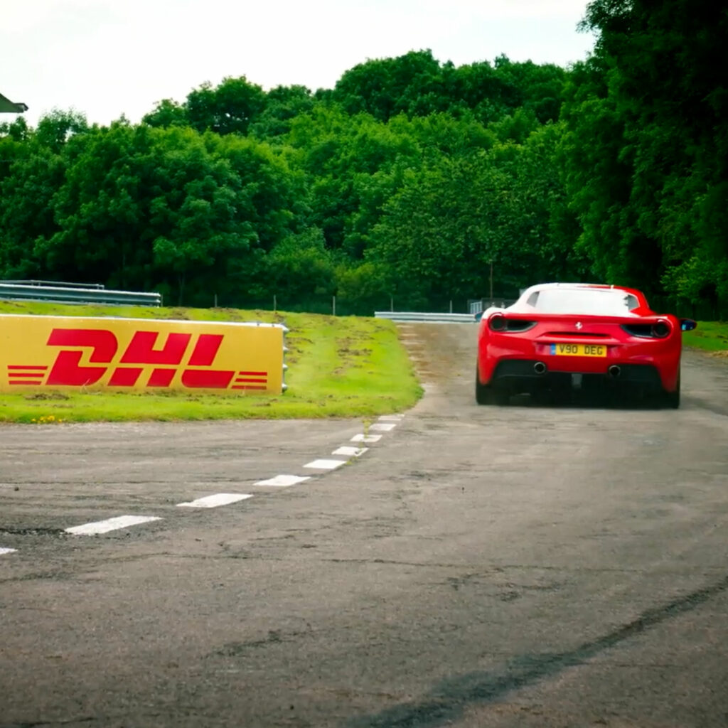 A red Ferrari on a race track passing a DHL-branded barrier.
