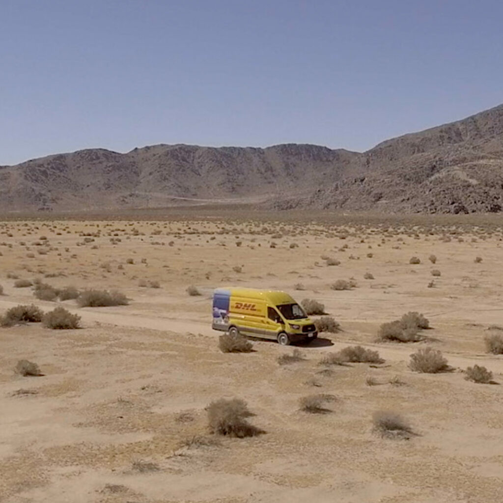 A DHL delivery van on a dirt road crossing open desert, with mountains on the horizon.