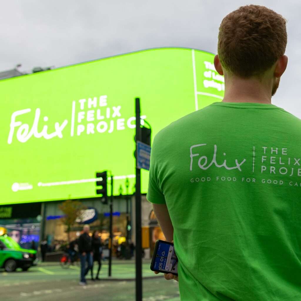 A Felix Project volunteer in a branded green T-shirt watches the charity's takeover of the Piccadilly Lights screen in London.