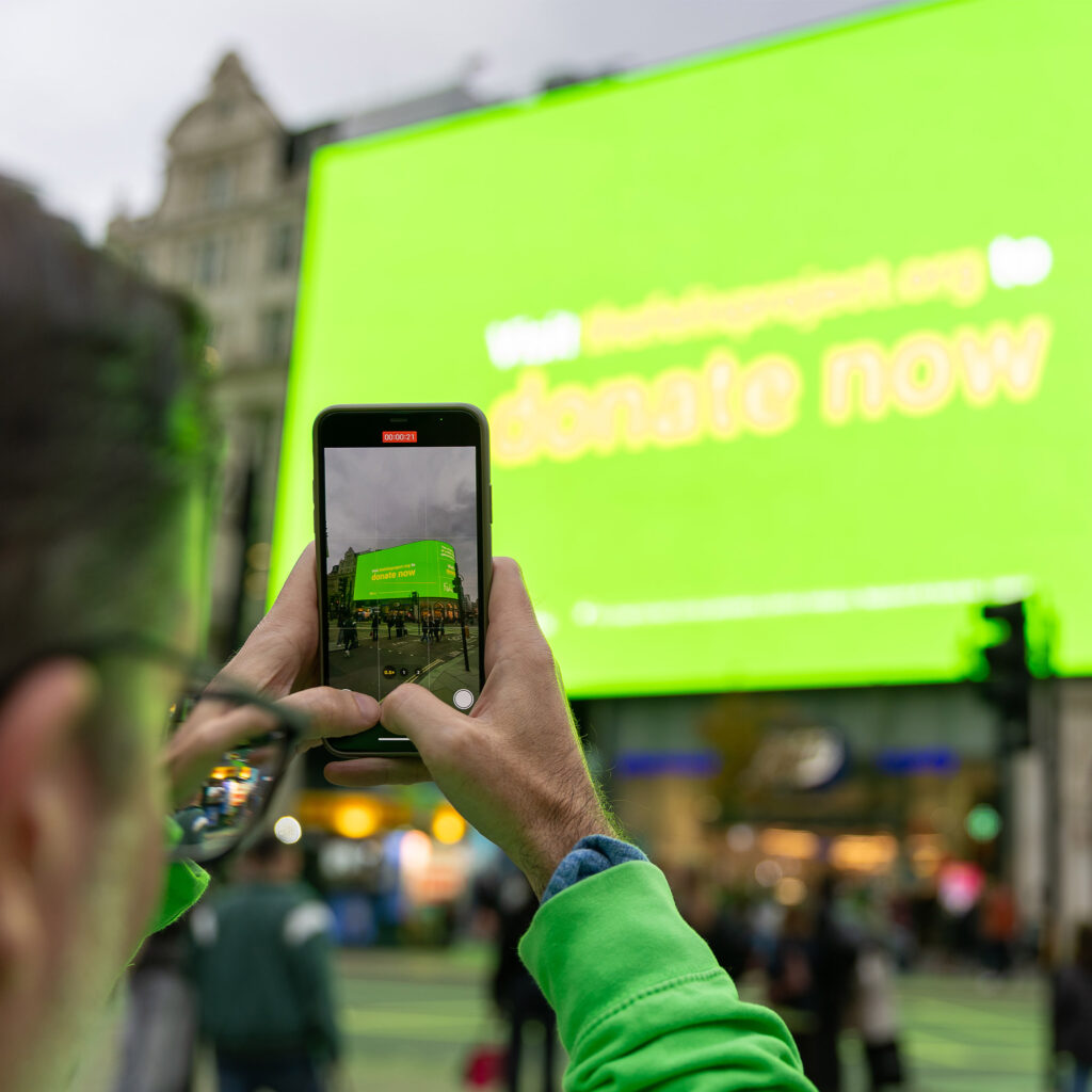 A passer-by films the Felix Project takeover on a phone as the Piccadilly Lights screen reads "Visit thefelixproject.org to donate now".