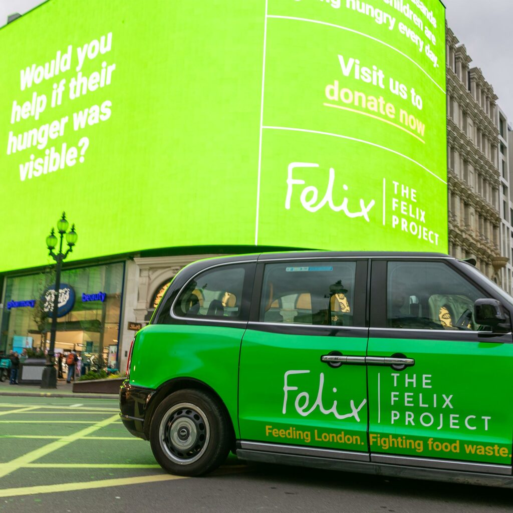 A green Felix Project-branded London taxi passes beneath the Piccadilly Lights screen asking "Would you help if their hunger was visible?"