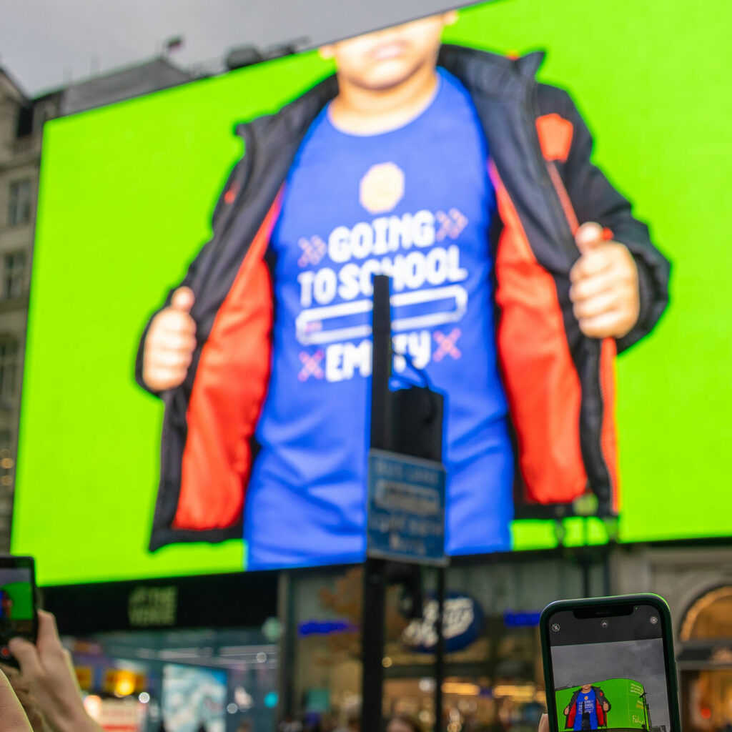 The Piccadilly Lights screen shows a child opening their coat to reveal a T-shirt reading "Going to school empty", as onlookers film on their phones.