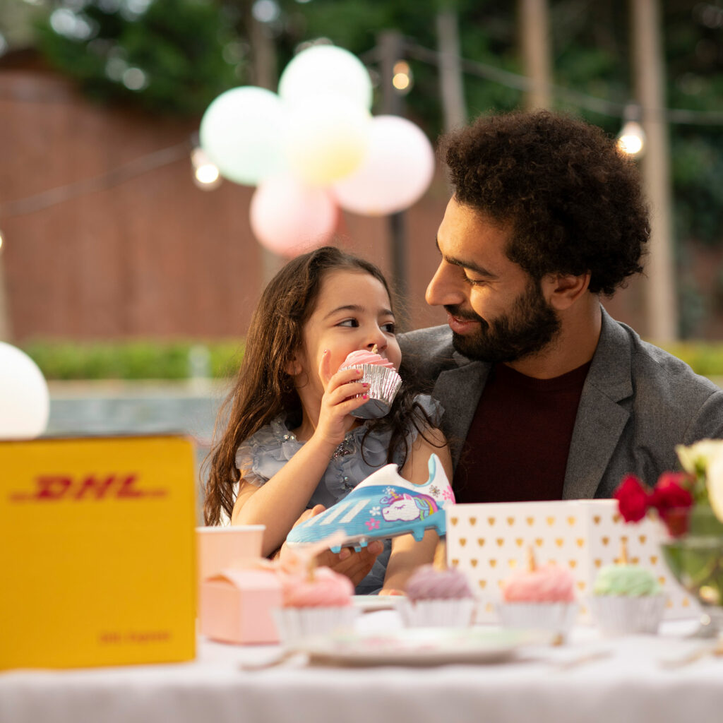 Mo Salah sits with a young girl at an outdoor party as she holds a unicorn-decorated football boot delivered in a DHL box.