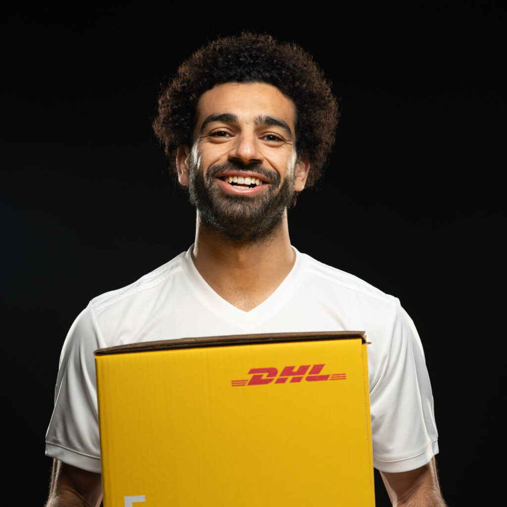 Studio portrait of Mo Salah in a white shirt, smiling and holding a yellow DHL box.