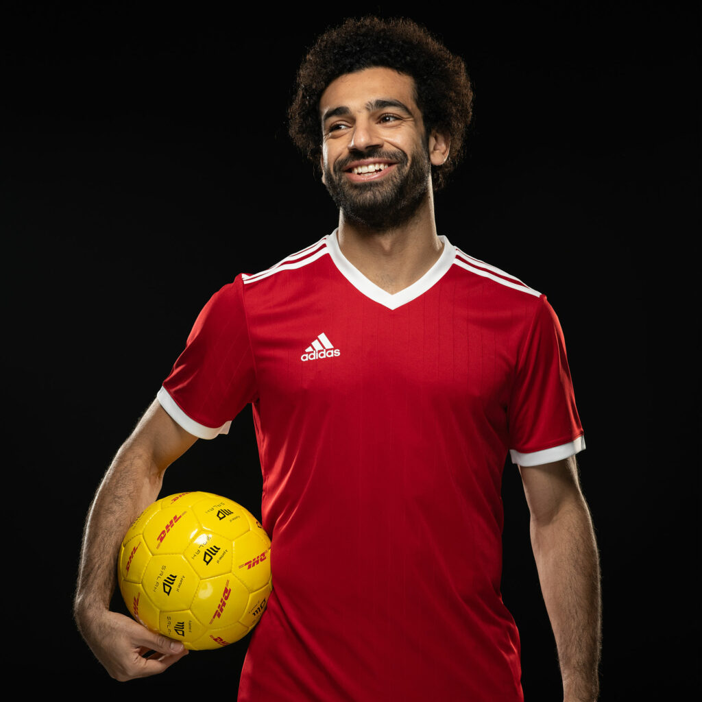 Studio portrait of Mo Salah in a red football shirt, holding a yellow ball branded with the DHL and Salah partnership logos.
