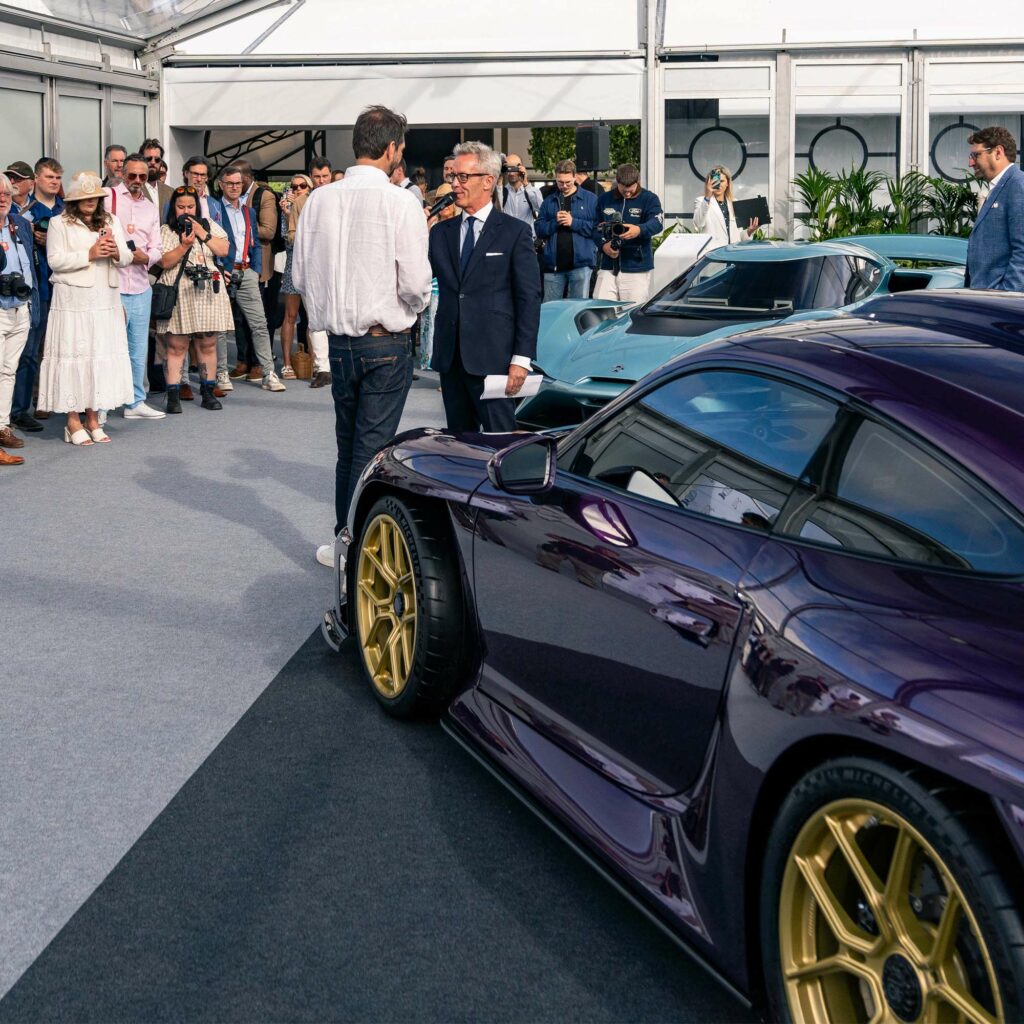 A presenter interviews a guest beside a purple supercar at Salon Prive, watched by guests and photographers.
