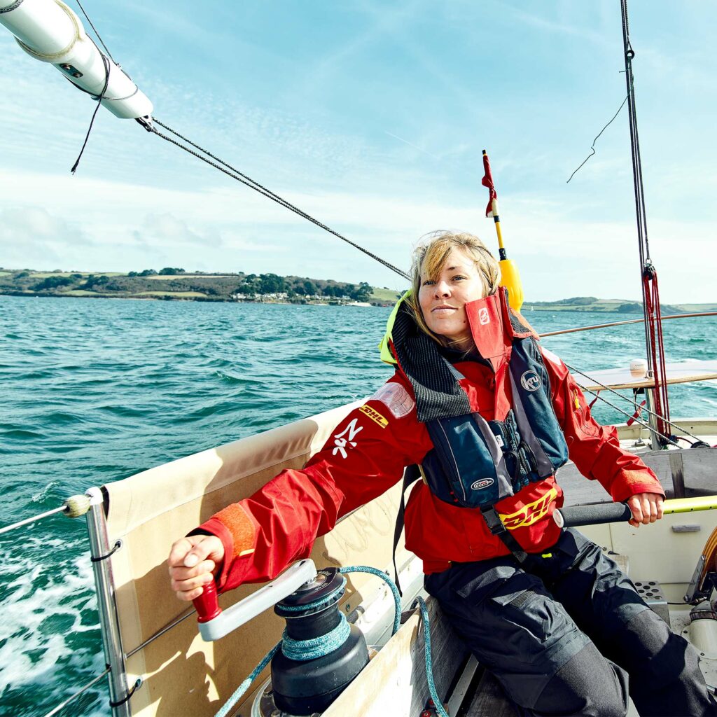 Susie Goodall seated in the cockpit working a winch handle, in DHL-branded foul weather gear, with a green coastline behind.