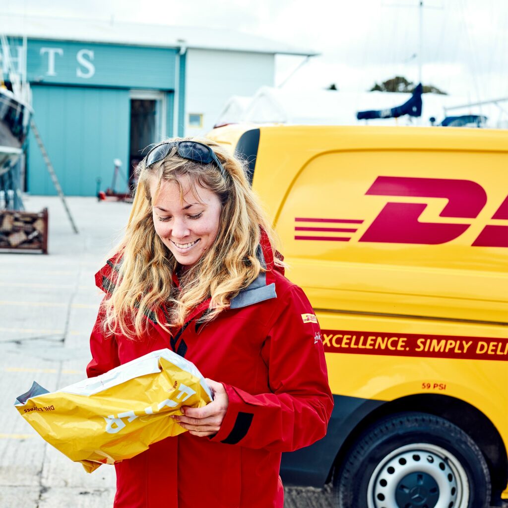 Susie Goodall smiling as she opens a DHL Express envelope beside a yellow DHL van carrying the line "Excellence. Simply delivered."