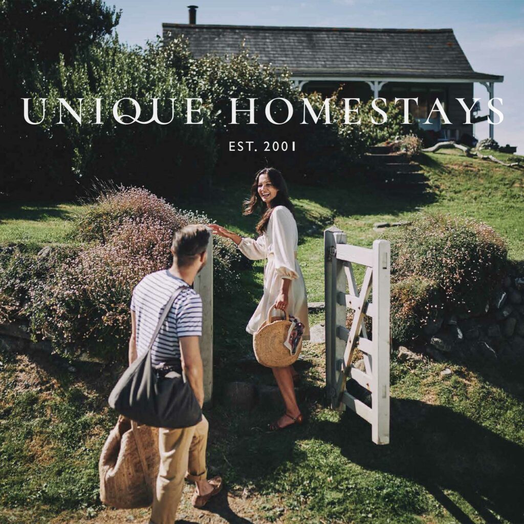 A couple arriving at a clifftop cottage, the woman turning back and reaching for her partner's hand, with the Unique Homestays wordmark across the frame.
