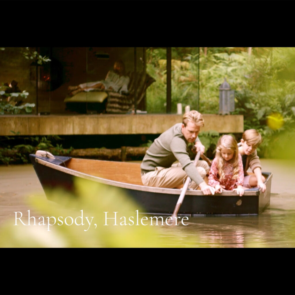 A still from the brand film captioned "Rhapsody, Haslemere", showing a father rowing two children across a lake beside a glass-fronted lodge.