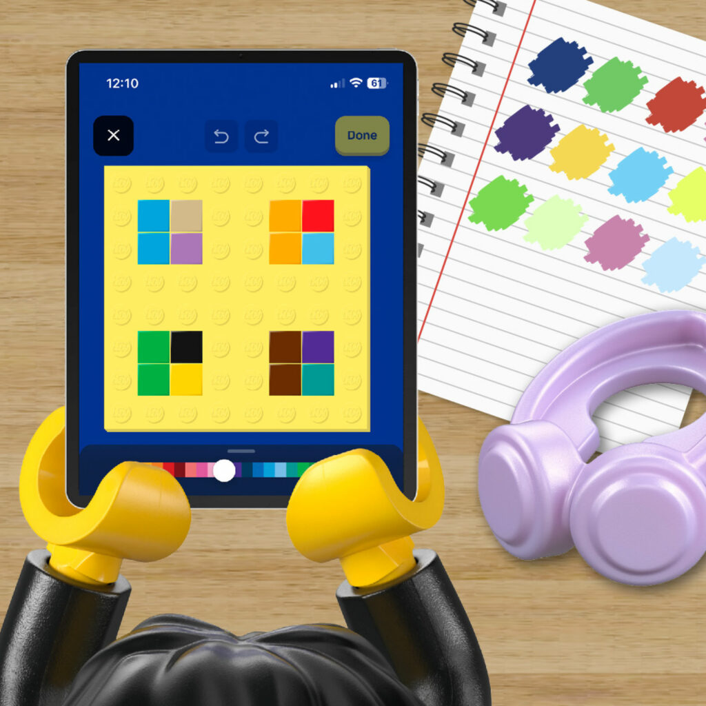 Ming holding a tablet showing a LEGO colour-picking app, beside a notebook of paint swatches and lilac headphones
